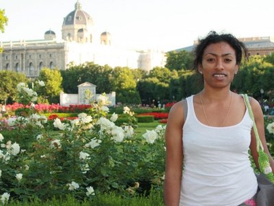 Kasha Tadel Gebre, in a white T-shirt, stands in front of a park with white flowers and a magnificent building in the background. 