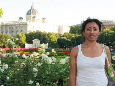 Kasha Tadel Gebre, in a white T-shirt, stands in front of a park with white flowers and a magnificent building in the background. 