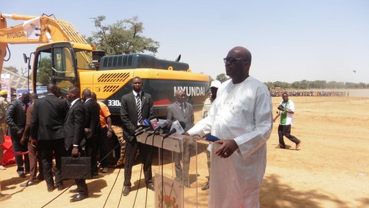 Man in long white dress standig in front of a group of man and a yellow  power shovel and behind a desk with five microphones on it