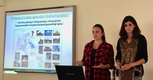 Two women are standing in front of their presentation on a screen, while one woman is talking 