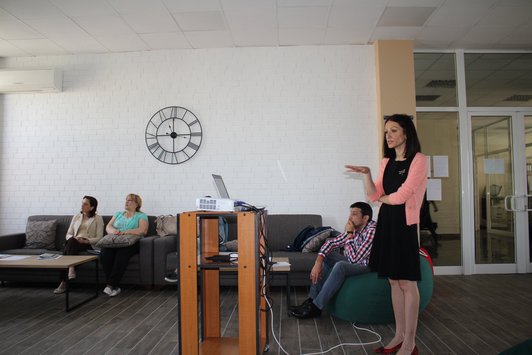 Woman standing behind a desk presenting somethin and other people are sitting and looking at the presentation in front of them