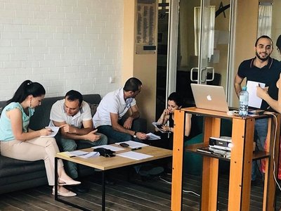 Some people sitting on a couch and some standing behind a desk with notebook on it and talking to each other