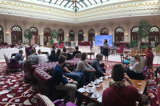 Reception hall with people sitting on couches around coffee tables with cups and small plates looking at a woman standing in front of a large flat screen giving a speech