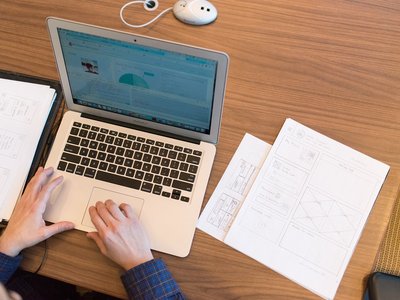 Aufgeklappter Laptop auf Tisch mit Händen an Tastatur, daneben weiße Notizblätter