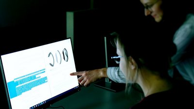 Zwei Schülerinnen sitzen vor einem Computerbildschirm. Eine davon zeigt auf das Display. 