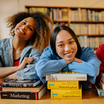 Drei Internationale Studierende sitzen in einer Bibliothek und stützen ihren Kopf auf Bücherstapeln auf.