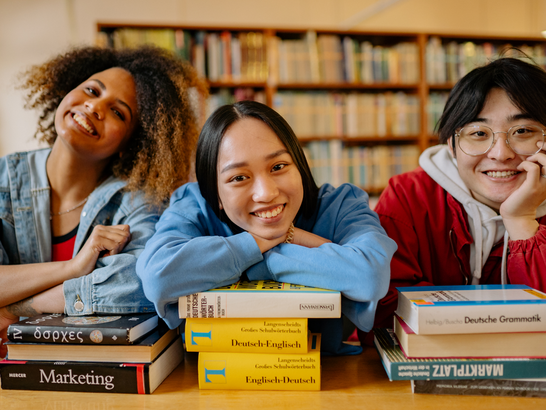 Drei Internationale Studierende sitzen in einer Bibliothek und stützen ihren Kopf auf Bücherstapeln auf.