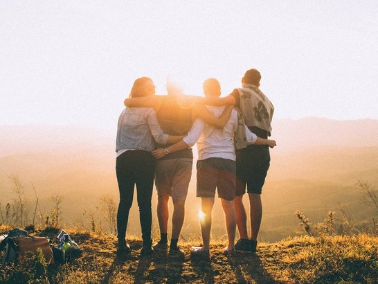  Four people put their arms around the shoulders or the person next to them. They stand at the top of a hill and watch the sunset.