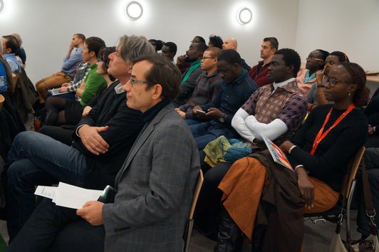 An audience sitting on chairs in a room with white a wall.