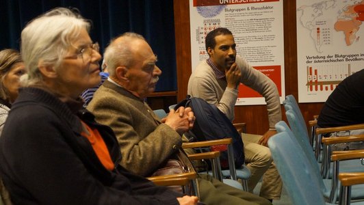 Six people as part of an audience, sitting on blue chairs