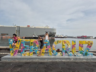 Drei Mädchen posieren an einem bunten, künstlerisch gestalteten Schriftzug 'VERACRUZ' am Hafen. Im Hintergrund liegt ein großes Frachtschiff mit dem Namen 'ADRIATIC HIGHWAY' im Wasser. Der Himmel ist leicht bewölkt, und weitere Schiffe sind in der Ferne zu sehen. Der Schriftzug ist farbenfroh mit folkloristischen Motiven bemalt.
