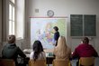 A classroom with a teacher in front of a world map and pupils looking in the teacher's direction.