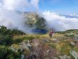  A young woman on a mountain can see a mountain lake diagonally below her and other hikers are coming behind her.