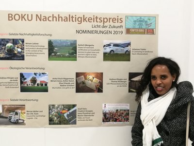 A woman standing in front of a large poster on a wall. The title on the poster reads "BOKU Nachhaltigkeitspreis - Licht der Zukunft - Nominierungen 2019"