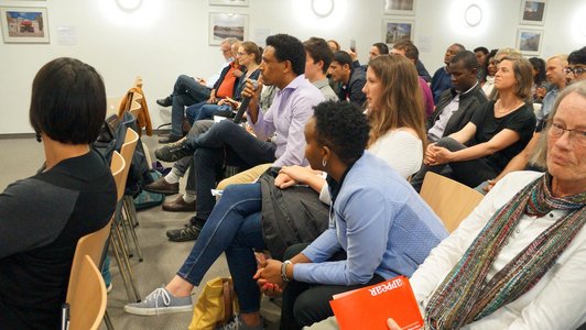 The picture shows an audience sitting on chairs. One man who is part of the audience speaks into a microphone.