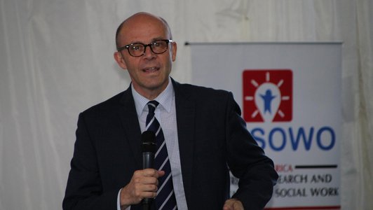 Man talking into a microphone in front of a conference flag