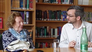 Contemporary witness Käthe Sasso (left in the picture) is talking to a man in his early 40s in a library room. There is a gas bottle of water on the table in front of them.