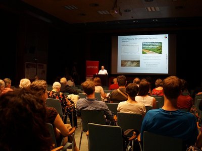 A man is delivering a speech from behind a speaker's desk in an auditorium. A presentation is projected onto the large screen behind him. The visible audience on the picture comprises approximately 30-35 people.