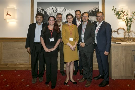 Two women and five men stand against a wall for a group photo and look in the same direction.