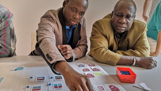 Two man sitting at a table putting blue coins on a poster to rate something