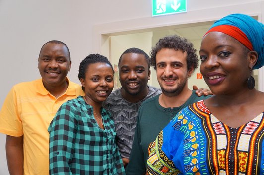 A group of five people is standing in a room and smiling into the camera.