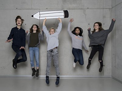 Five children jump up, one holds a large cardboard pencil in his hands.