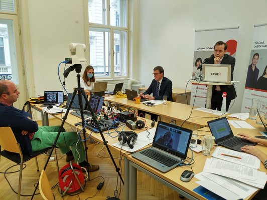 Table with three speakers, two in front of it sitting in front of a laptop, one standing at the desk, and a technician sitting in front of a control panel and several screens