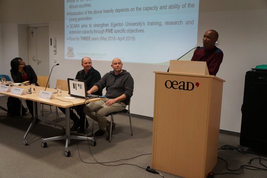 Three people are sitting at a table with name plates, glasses of water, sheets of paper and a laptop on it while one person is giving a speech at a speaker's desk next to them.