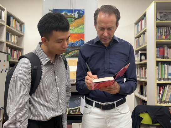 Ein junger und ein älterer Mann, im HIntergrund eine Bibliothek. Der ältere Mann hält ein Buch in der Hand und signiert es