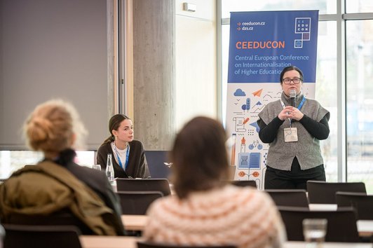 die Frau mit Mikro präsentiert; die andere zuhört; die Leuten sitzen im Saal in Hintergrund 