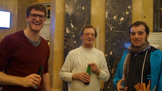 Three men are standing around a table in a room with marble walls, holding drinks in their hands.