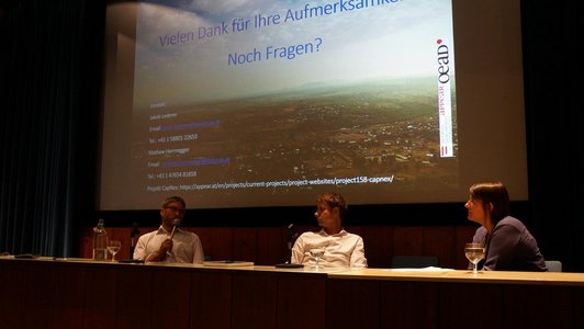 One woman and two men are sitting at a long desk with glasses and notebooks on it. One of the men is holding a microphone and speaking to the others. A presentation is projected onto the screen behind them. 