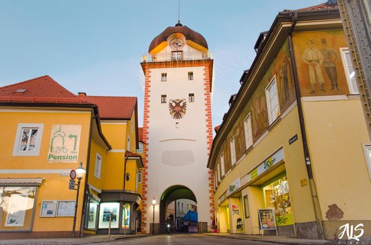 Ein Turm mit einem offenen Tor an seinem Fuß und einer Uhr. Er ist inmitten einer Gasse.