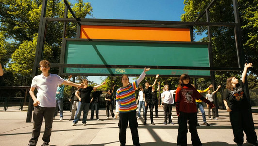 Junge Menschen winken vor einem großen Metallkonstrukt.