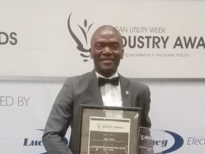 A man who has just received the African Utility Week Industry Award, standing in front of a large roll-up announcing the event.