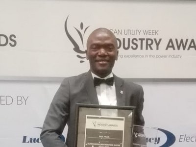 A man who has just received the African Utility Week Industry Award, standing in front of a large roll-up announcing the event.