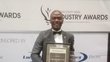 A man who has just received the African Utility Week Industry Award, standing in front of a large roll-up announcing the event.