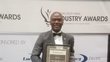 A man who has just received the African Utility Week Industry Award, standing in front of a large roll-up announcing the event.