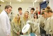 Students on the AT&S company tour in Shanghai. They wear lab coats and safety goggles and look at a large hanging magnifying glass with circuit boards underneath.