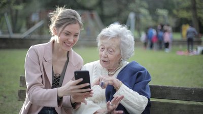 Eine junge weiße Frau zeigt einer älteren weißen Dame ihren Smartphone. Die junge Dame lächelt und die ältere Dame wirkt positiv überrascht.