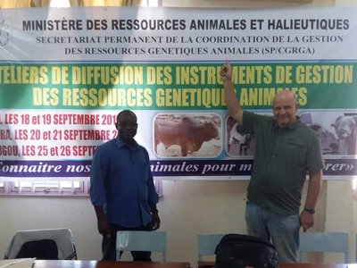 Two man smiling and standing in front of a big poster of the ministry, while one man is ponting at the poster