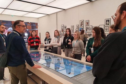 Menschen um einen rechteckigen Tisch mit Bildschirm, im Hintergrund eine weiße Wand mit vielen gerahmten Fotos