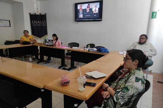 Ein Klassenraum mit Tischen in O-Form, runderhum 4 Personen, im Hintergrund ein Bildschirm mit Videokonferenz