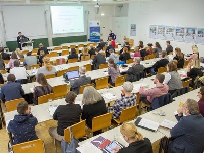 Viele Personen sitzen an Tischreihen in einem Seminarraum und schauen nach vorne zu einem Mann, der am Podium steht und einen Laptop auf einem Stehtisch vor sich stehen hat und zum Publikum schaut