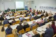 Viele Personen sitzen an Tischreihen in einem Seminarraum und schauen nach vorne zu einem Mann, der am Podium steht und einen Laptop auf einem Stehtisch vor sich stehen hat und zum Publikum schaut