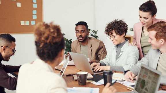 Ein Team unterhält sich im Büro. Das Team besteht aus mehreren jungen Menschen mit Laptops. Alle sitzen an einem Holz Tisch.