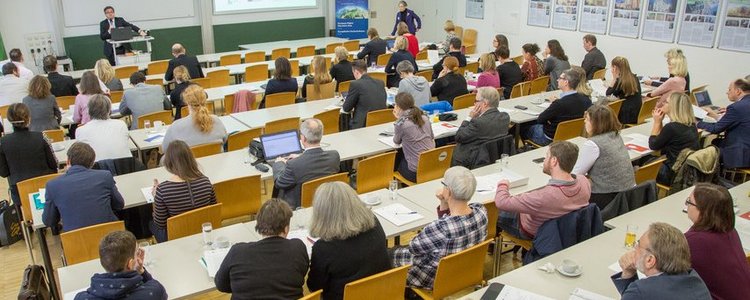 Viele Personen sitzen an Tischreihen in einem Seminarraum und schauen nach vorne zu einem Mann, der am Podium steht und einen Laptop auf einem Stehtisch vor sich stehen hat und zum Publikum schaut