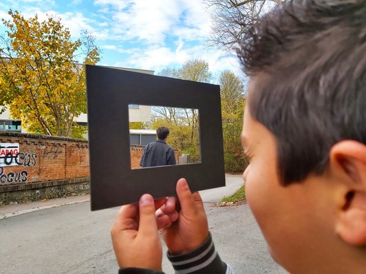 Ein Junge hält einen leeren Rahmen hoch, durch das Loch ist ein anderer junger Mensch zu sehen.