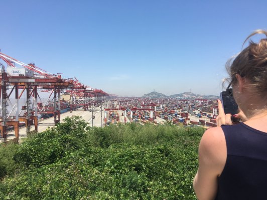  Student takes a photo of the deep-sea port of Shanghai with her smartphone. Directly in front of her is a garden full of green plants, behind her is the port with cranes and containers and in the distance some mountains