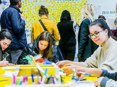 Young people doing crafts in class. They are sitting on a yellow table. In the background is a wall with favorite words.